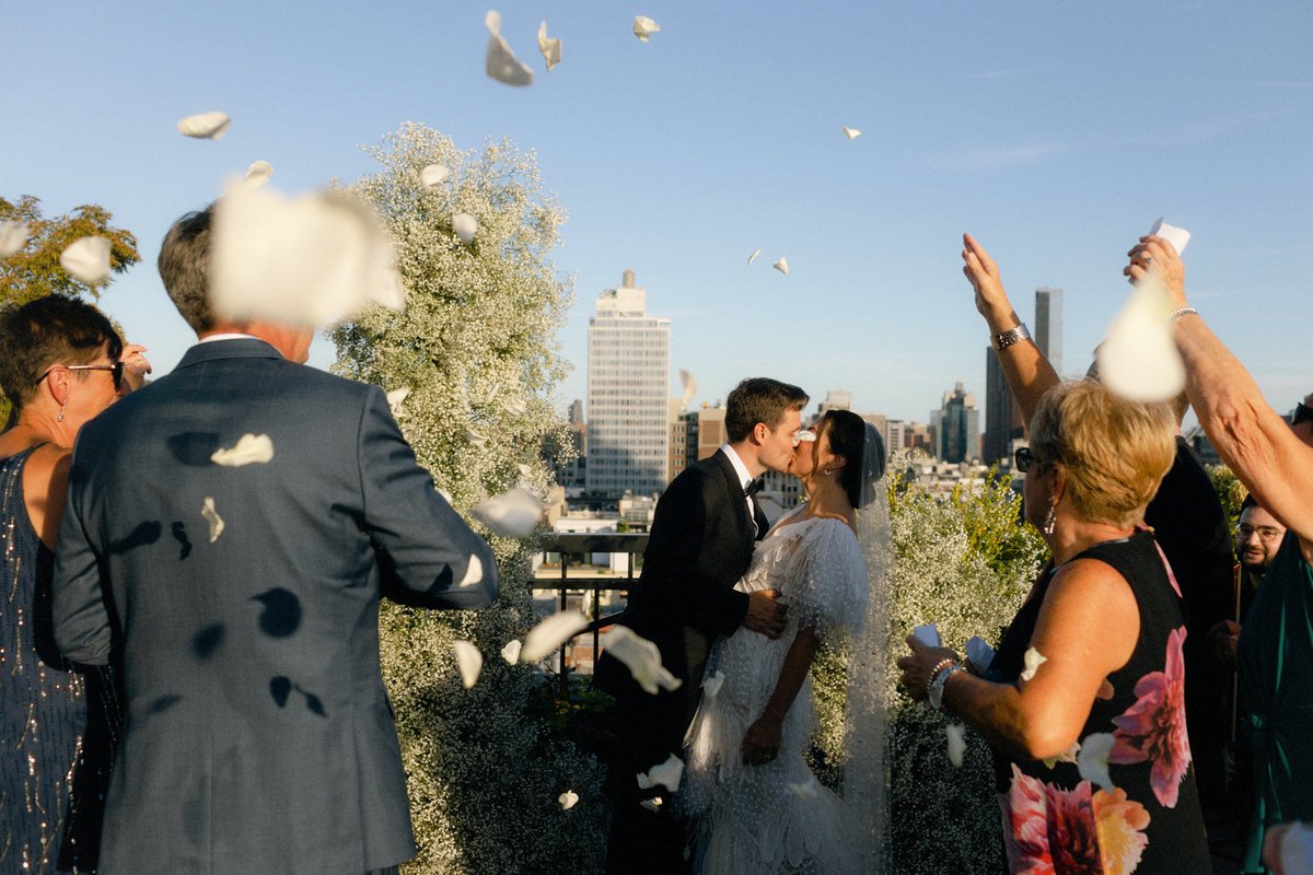 wedding guests throwing white petals while the newlyweds kiss on a Manhattan rooftop