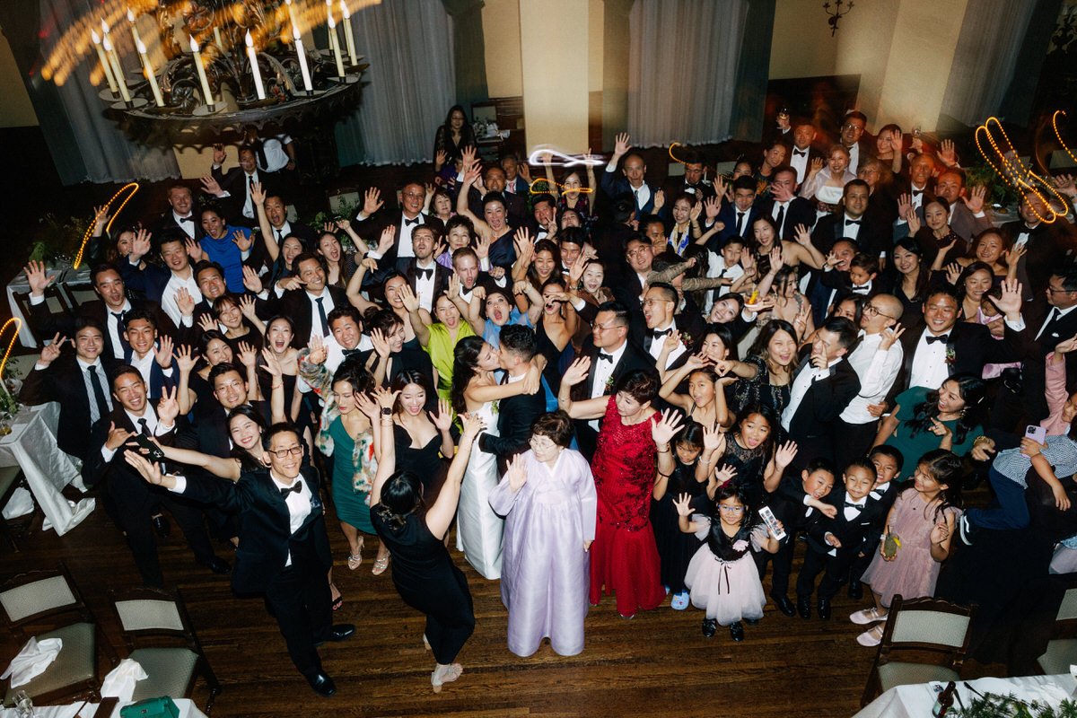 large group photo of wedding guests celebrating while bride and groom kiss in centre