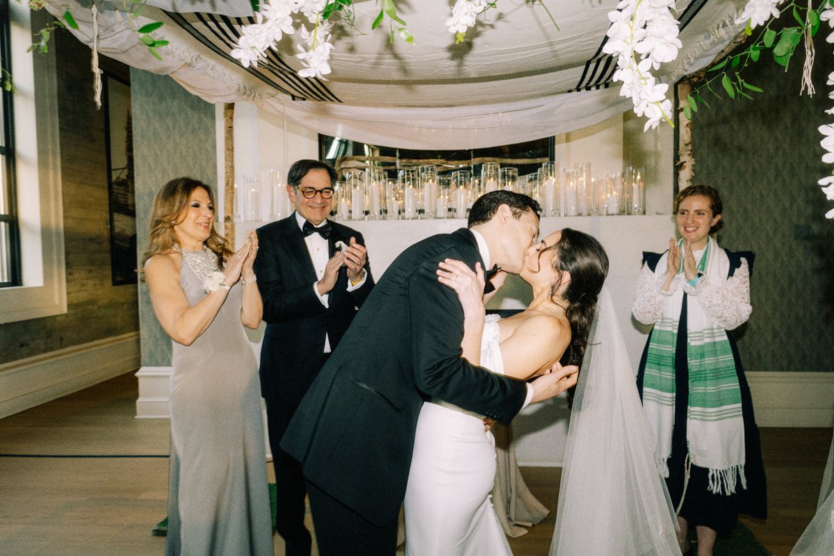 wedding couple kissing under the Chuppah while their family and Rabbi surround them at the Switch House by Cescaphe