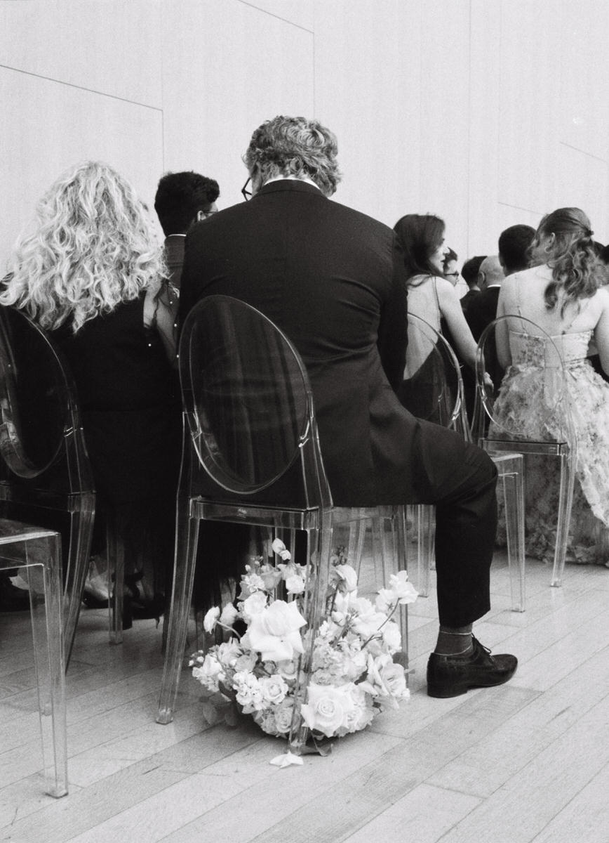 wedding guest sitting on a clear chair with a floral arrangement underneath it