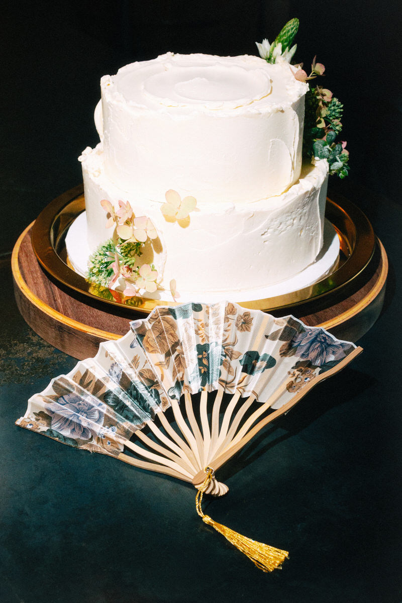 simple vanilla cream wedding cake featuring two tiers and hydrangea petals. the cake is resting on a copper plate and wooden stand, stylized with a floral hand fan