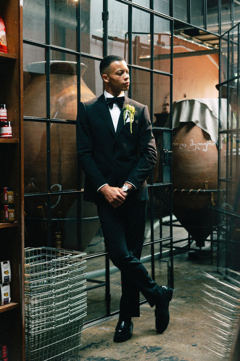 handsome biracial groom in emporio armani posing in front of a glass partition wall inside la fabrique st george winery in vancouver